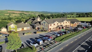 A hotel with a parking lot, a road in the foreground, and a green landscape in the background. - Rhu Glenn (Waterford)