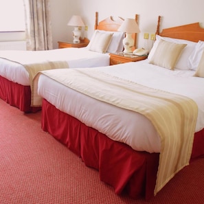 Habitación de hotel con dos camas, cada una con cabecero de madera, ropa de cama blanca y falda roja. Hay mesitas de noche con lámparas y una ventana con cortinas.