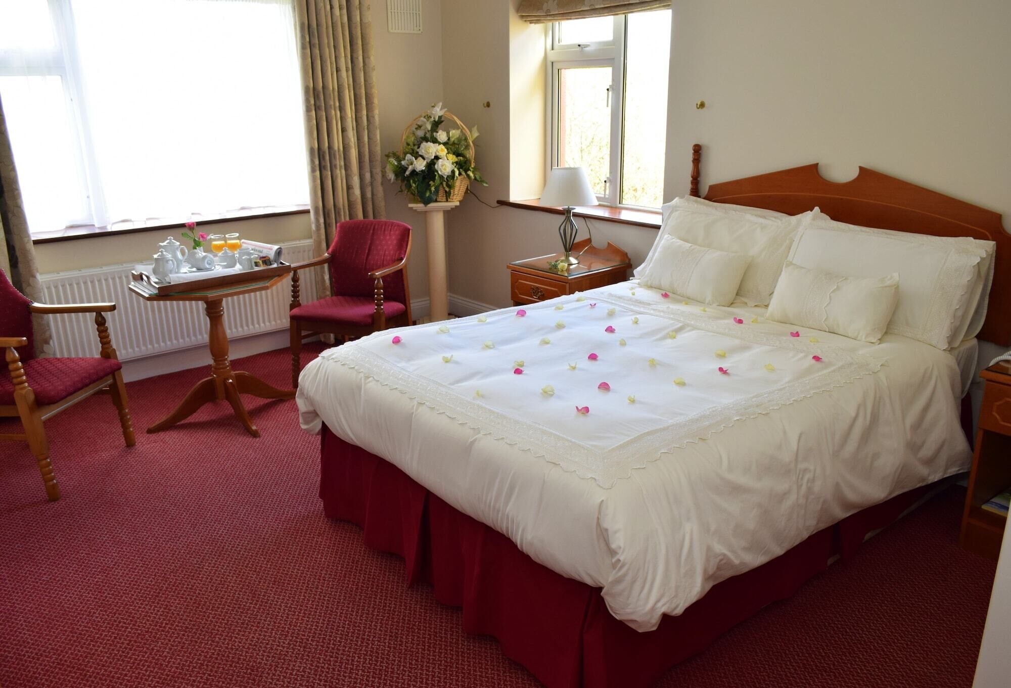 A hotel room with a large bed, a desk with a chair, a small table with a basket of flowers, and a window with curtains.
