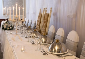 A banquet table set with silverware, glasses, and covered dishes, with candles and a floral arrangement.