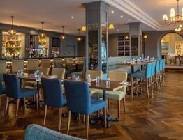 Un restaurante con piso de madera, sillas azules y beige, y una zona de bar con una exhibición de licores visible.