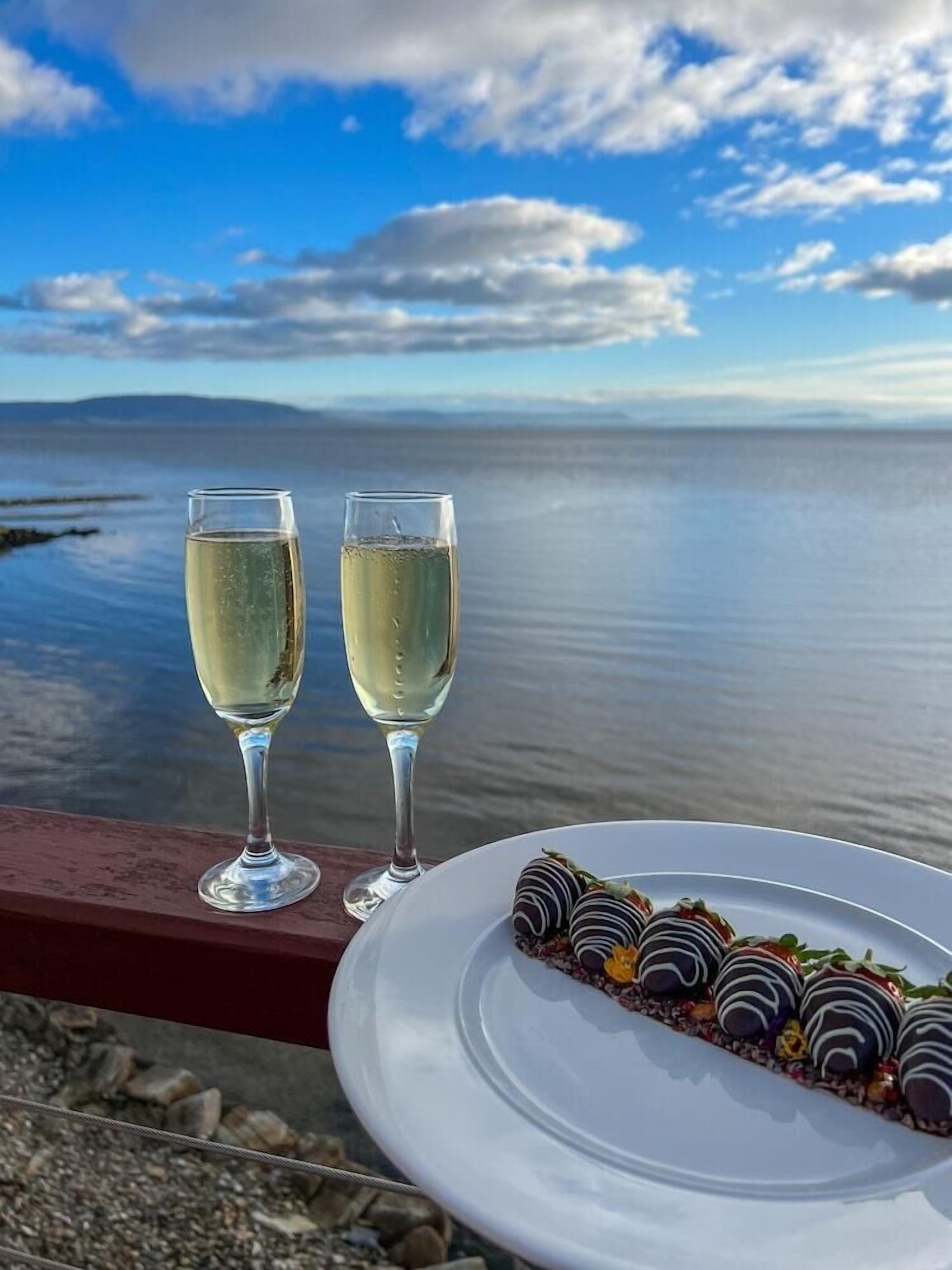 Two glasses of white wine on a balcony overlooking a calm body of water with a clear sky.