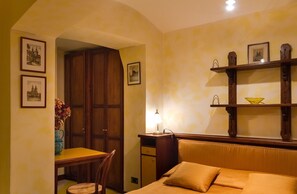 Una habitación de hotel con cama, escritorio, silla, lámpara y estante con un bol.