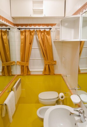 Un baño con paredes amarillas, un lavamanos blanco, dos inodoros y cortinas naranjas.