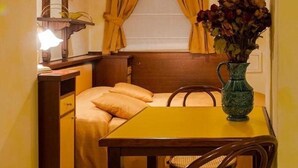 A compact room with a bed, a small table, and a chair. There is a window with curtains, a shelf with books, and a vase with flowers. - Residence Tynska (Prague)