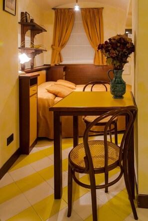 Una habitación compacta con una cama, una mesita con un florero, una silla y una lámpara.