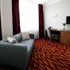 A hotel room with a grey sofa, a wooden desk, a chair, a TV, and a patterned carpet. - Hotel Central (Plzen)