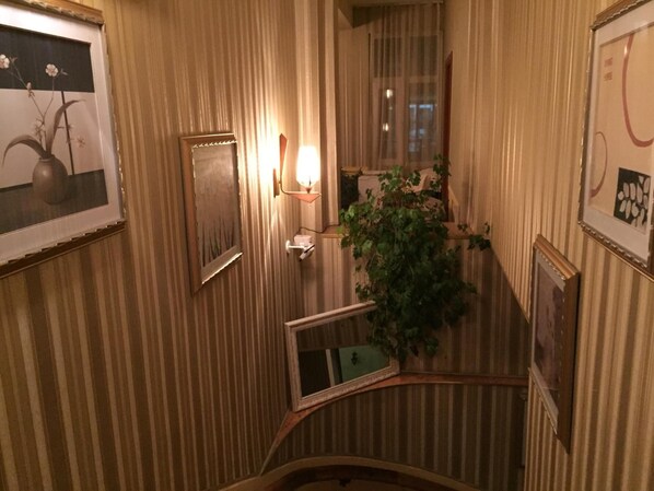 A narrow hallway with striped wallpaper, a framed picture on the left, a wall-mounted light fixture, and a potted plant. - Chipilakoff (Burgas)