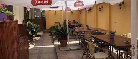 An outdoor dining area with tables and chairs, potted plants, and umbrellas with signage.