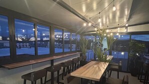 An outdoor dining area with a long wooden table and chairs, large windows, and string lights. - City Hotel (Veliko Tarnovo)