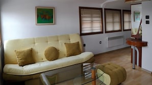 A living room with a beige sofa, a glass coffee table, and a wooden side table. There are three windows with blinds, a radiator, and a painting on the wall. - Gurko (Veliko Tarnovo)