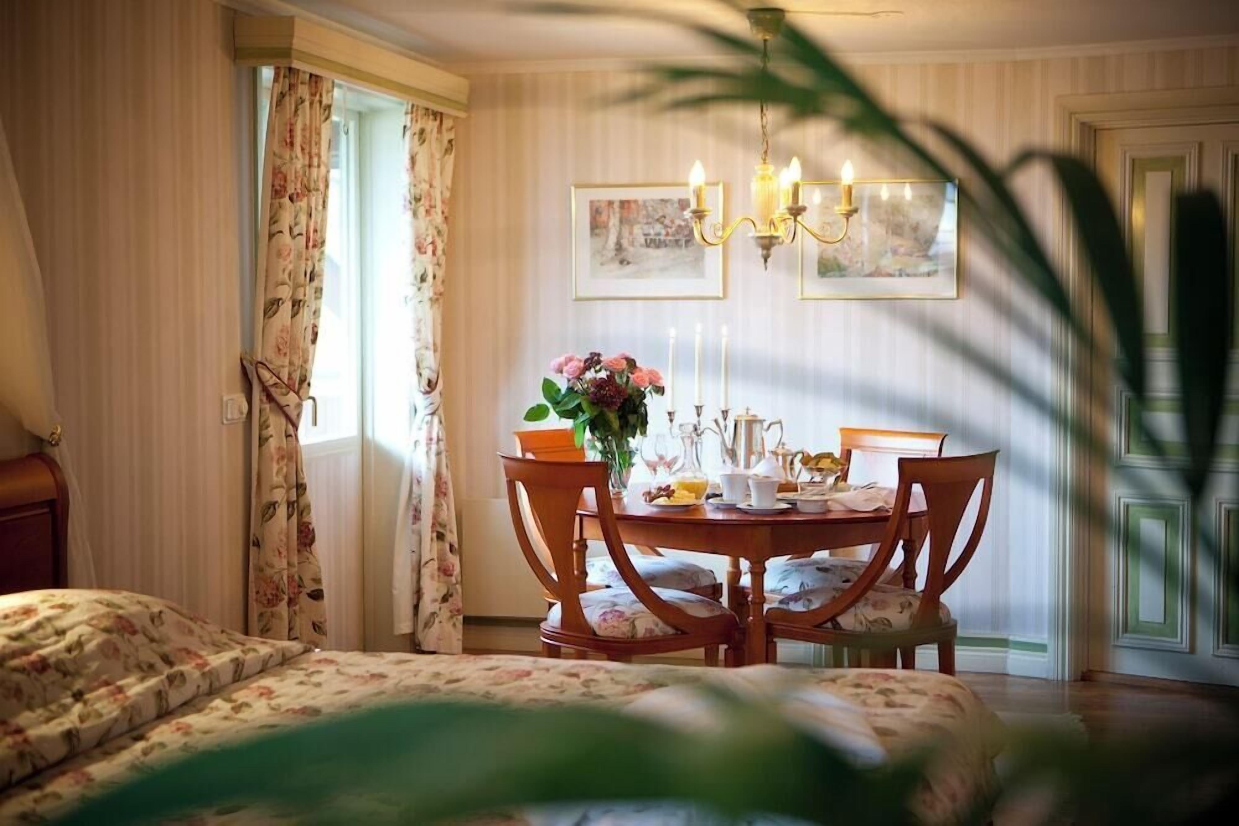 A room with a bed, a dining table with chairs, a chandelier, and a window with curtains.