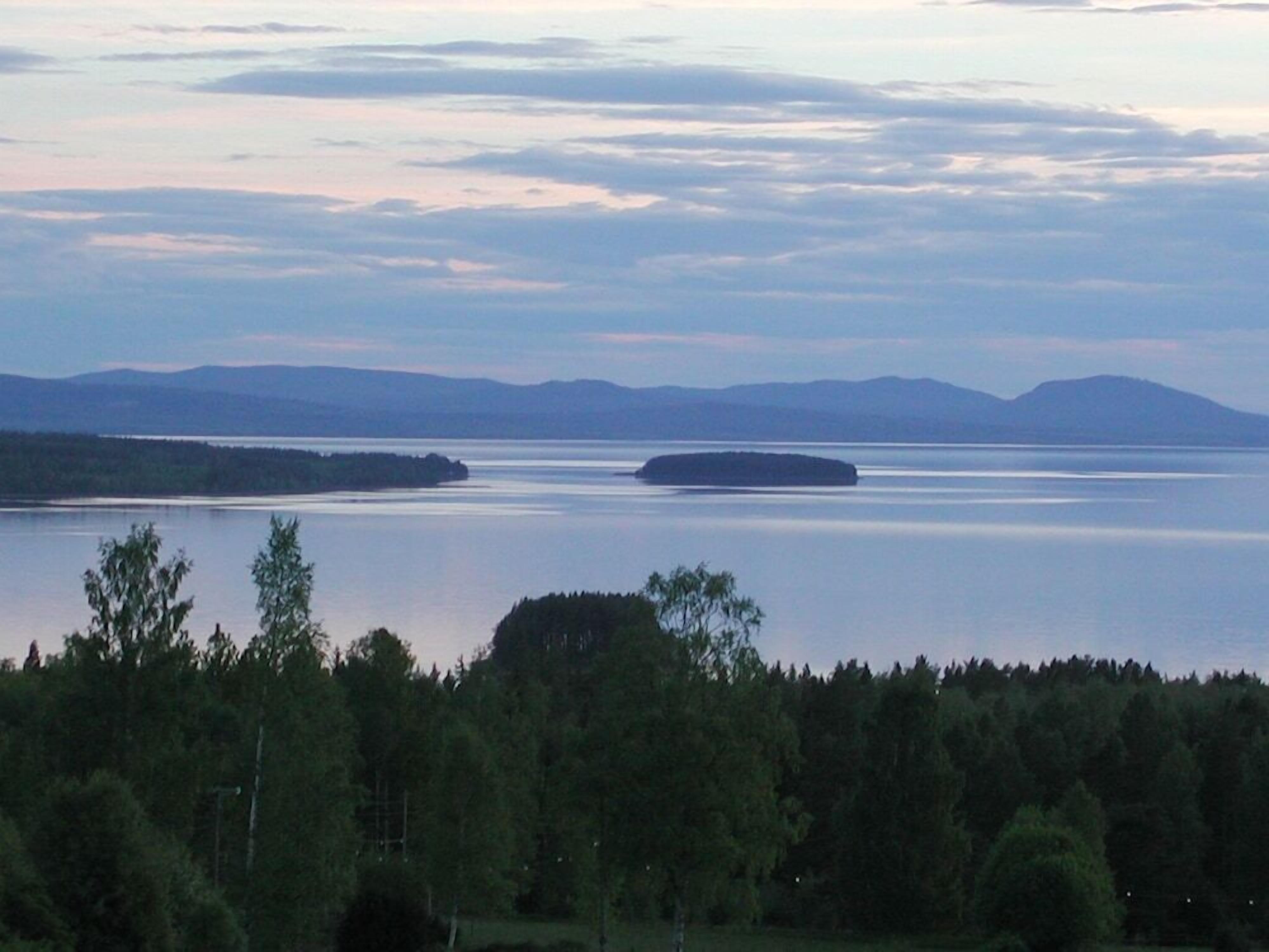 A large body of water with a small island in the center, surrounded by forested land and distant mountains.