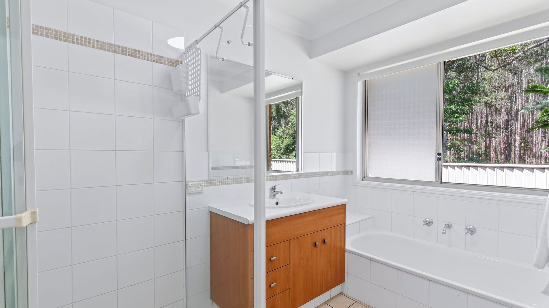 Un bagno moderno rivestito di piastrelle bianche, con un mobile vanitario in legno, uno specchio grande, una vasca da bagno e una finestra con vista sugli alberi.