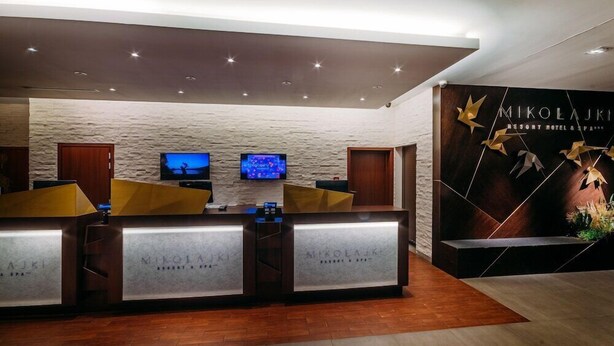A reception area with a stone wall backdrop, two reception desks, and a large poster advertising the resort. - Hotel Mikolajki Resort By Desilva (Mikolajki)