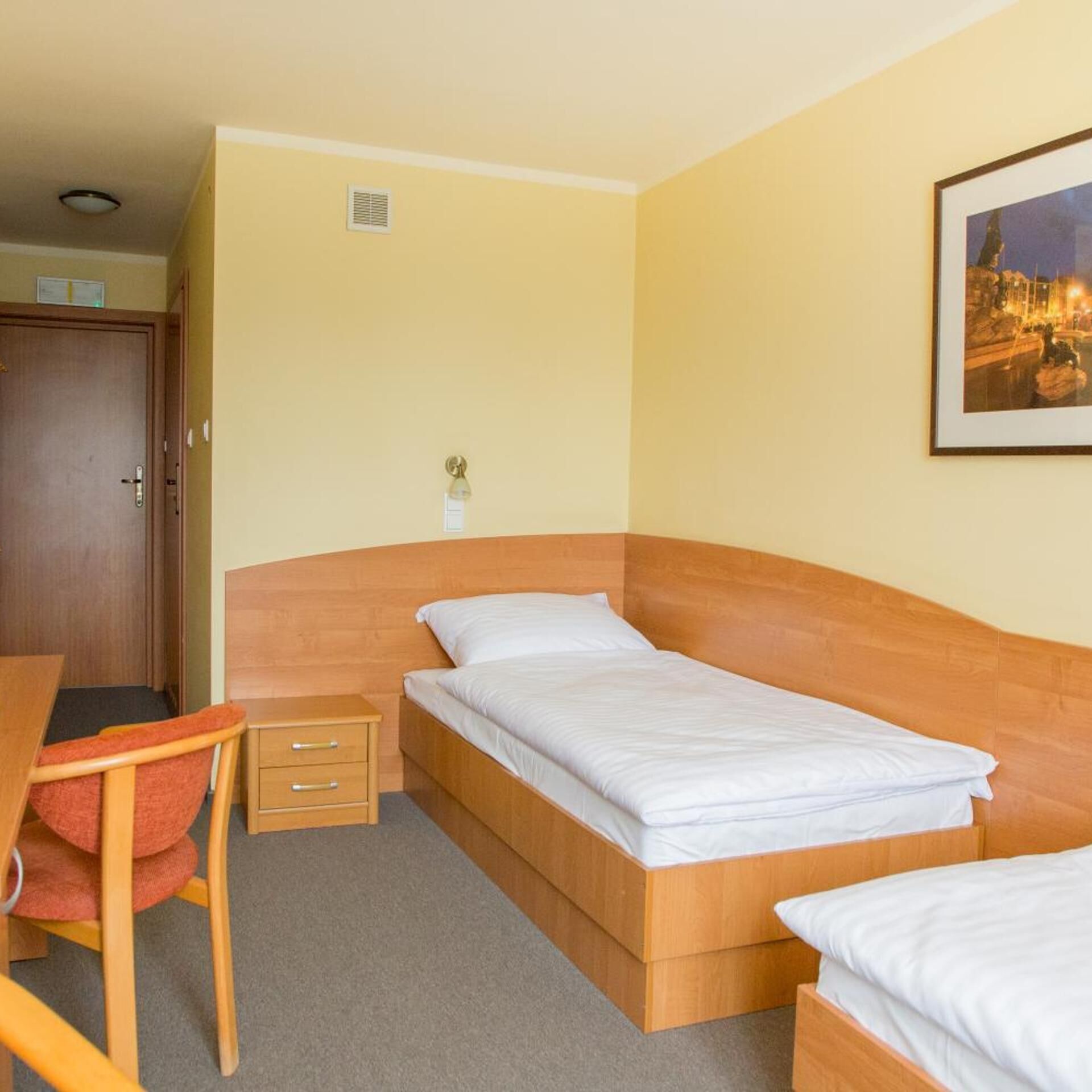 A single bed with a wooden headboard, a bedside table with drawers, a chair, and a framed picture on the wall.