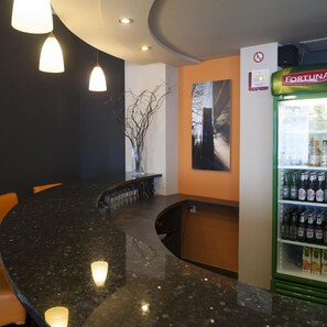 Zona de bar con mostrador curvo de granito, lámparas colgantes, una obra de arte en la pared y un refrigerador lleno de bebidas.