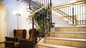 Una escalera con barandilla de metal, escalones alfombrados y un sillón marrón en una habitación con pared blanca y una planta.