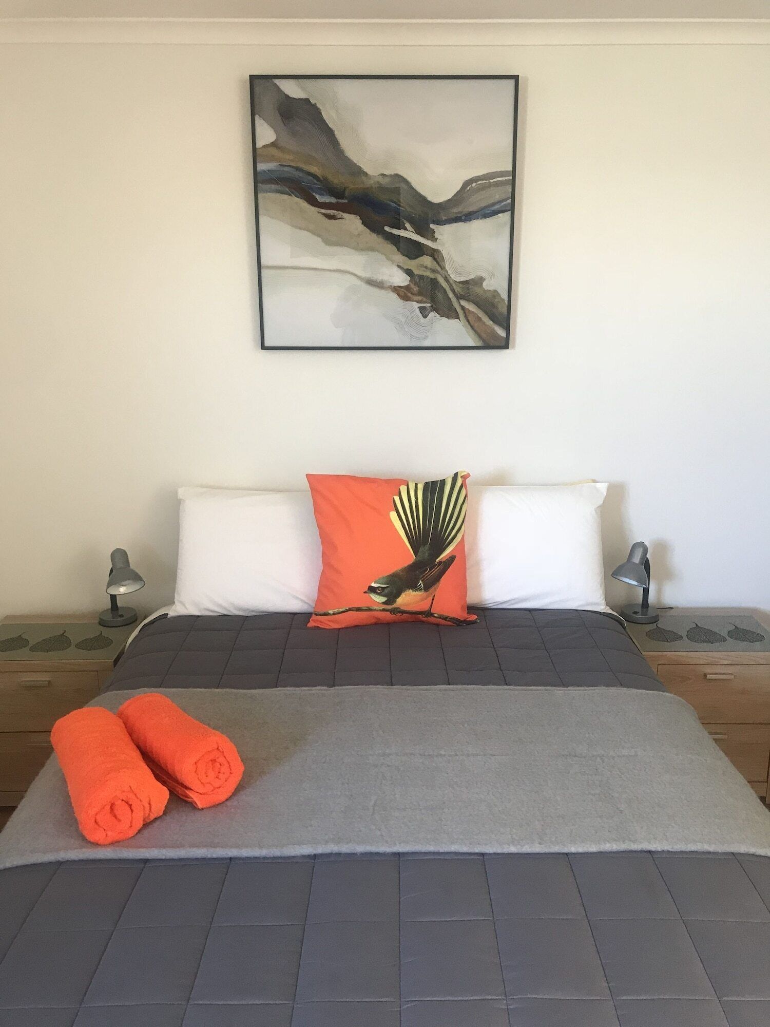 Ein ordentlich bezogenes Bett mit einer grauen Tagesdecke, zwei weißen Kissen und einem orangefarbenen Kissen mit Vogelmotiv. Darüber hängt ein gerahmtes abstraktes Gemälde, und auf den hölzernen Nachttischen stehen jeweils eine Nachttischlampe.