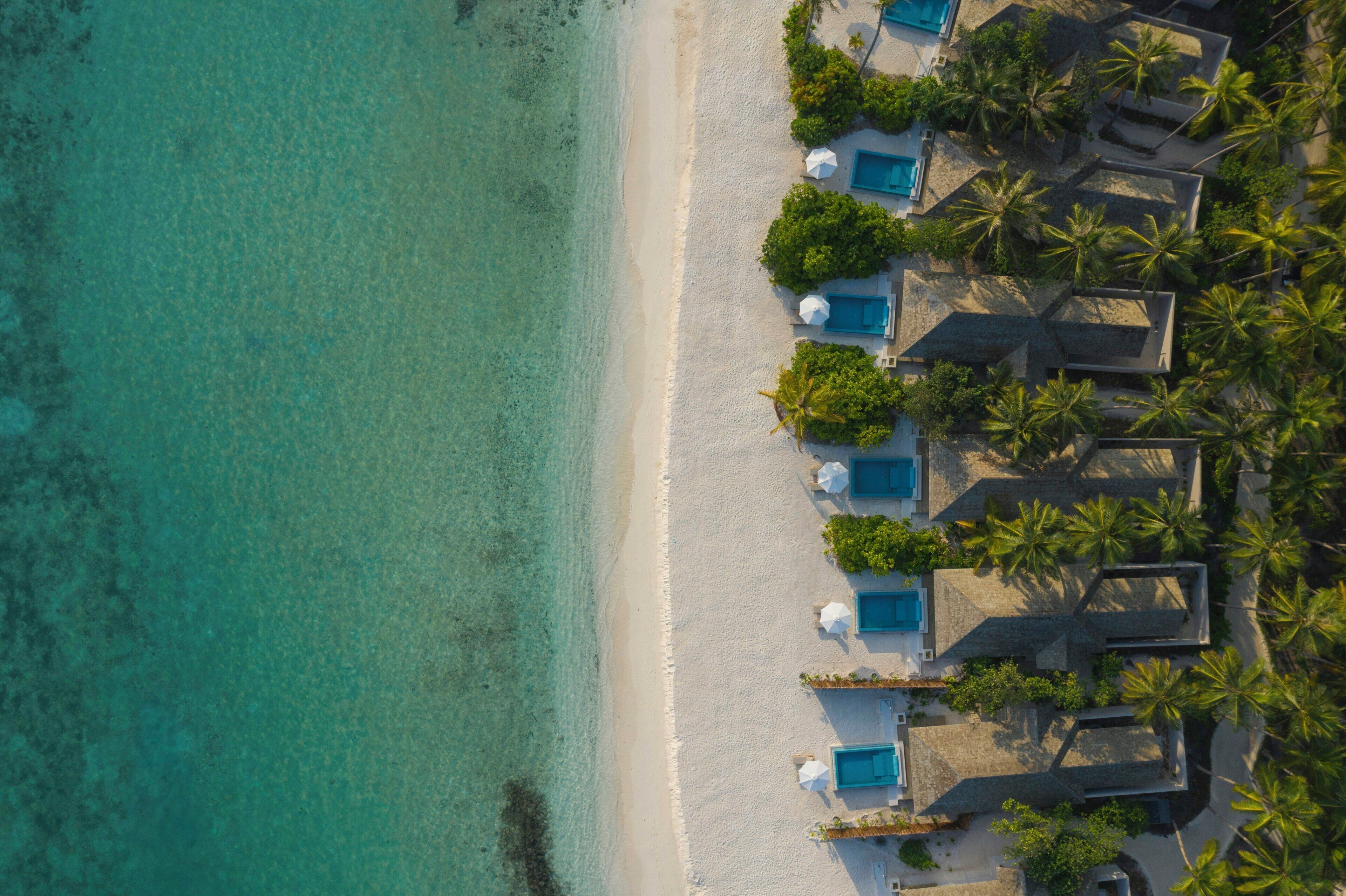 Vista aérea de un resort en la playa con varias estructuras de techo azul, palmeras y agua turquesa cristalina.