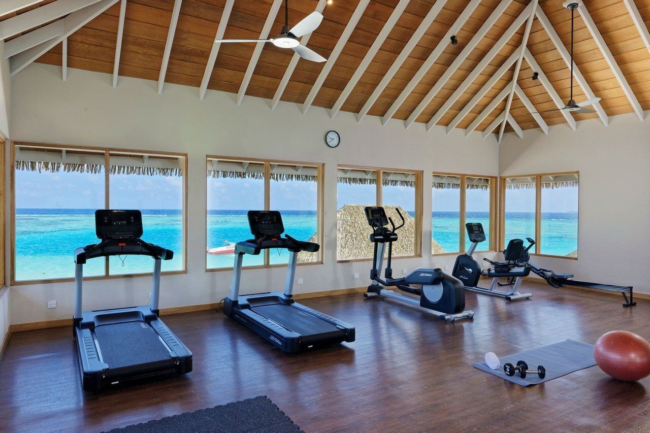 A gym with treadmills, an elliptical, and a rowing machine, large windows with ocean views, and a wooden ceiling.