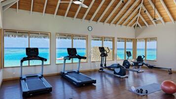 A gym with treadmills, an elliptical, and a rowing machine, large windows with ocean views, and a wooden ceiling.