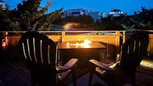 A wooden deck with a table and chairs, a fire feature, and houses in the background. - 7221 - Wind & Waves Bungalow - Salvo Beach Bungalow w/ Pool & Hot Tub (Salvo)