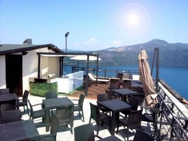 Terraza al aire libre con mesas y sillas, una pequeña zona de bar y vistas a un lago y montañas.