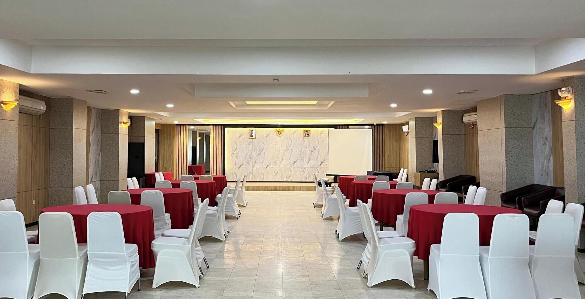 A conference room with rows of tables and chairs, a projector screen, and a marble wall.