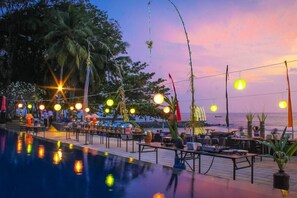 An outdoor pool area with tables and chairs, surrounded by palm trees and a sunset view. - Mambruk Anyer (Anyer)