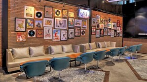 A lounge area with a long sofa and multiple chairs, a brick wall with framed posters and vinyl records, and a modern ceiling design. - Mambruk Anyer (Anyer)