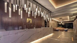 A modern lobby with a marble reception desk, hanging pendant lights, and a seating area with sofas and ottomans. - Mambruk Anyer (Anyer)