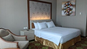 A hotel room with a bed, a nightstand, a chair, a headboard with a light, and a wall art piece.