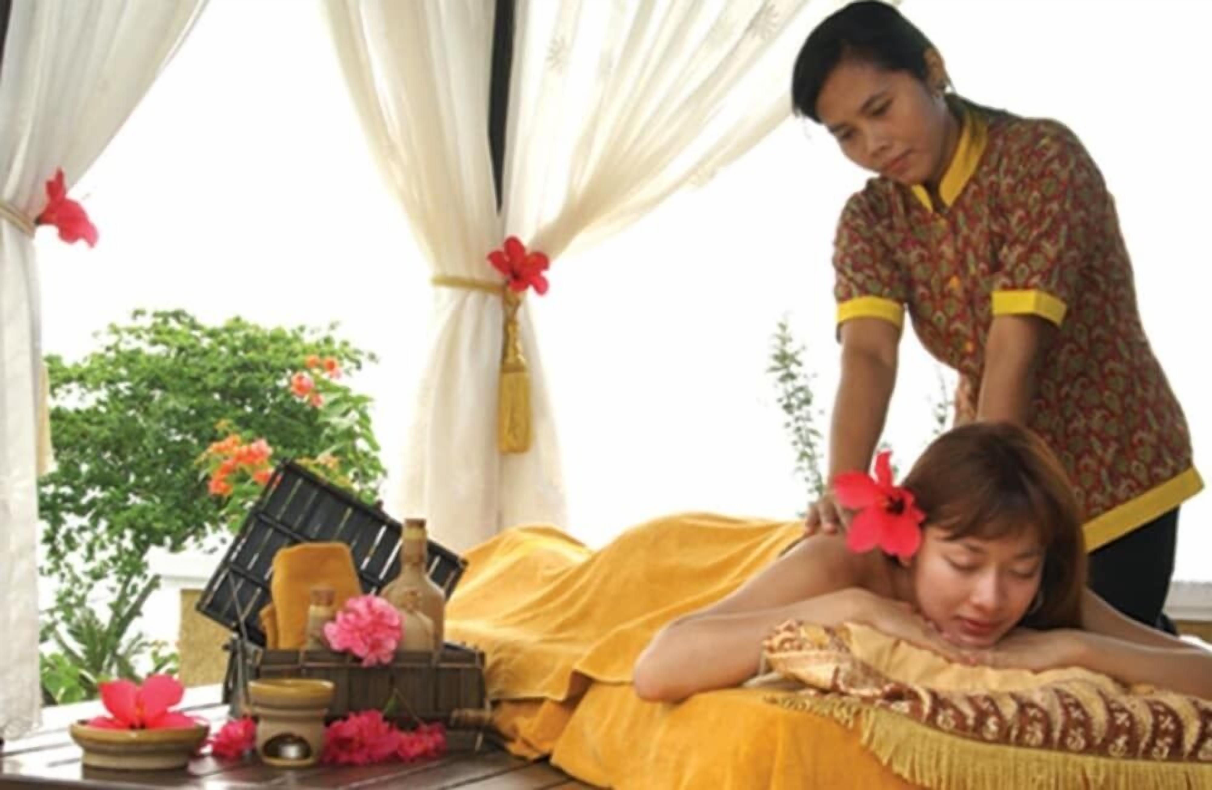 A massage therapy session in a room with a bed, a massage table, and a window with curtains.