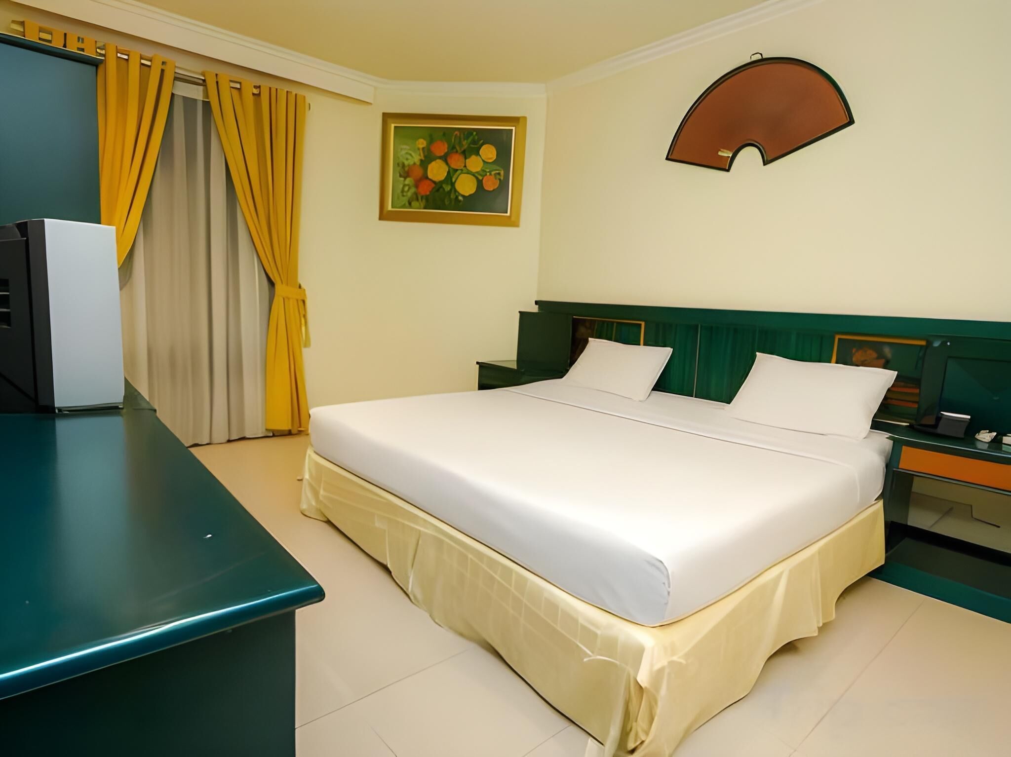 Une chambre d’hôtel avec un lit, un bureau, une chaise, une table de chevet, une lampe, un tableau et une fenêtre avec des rideaux.