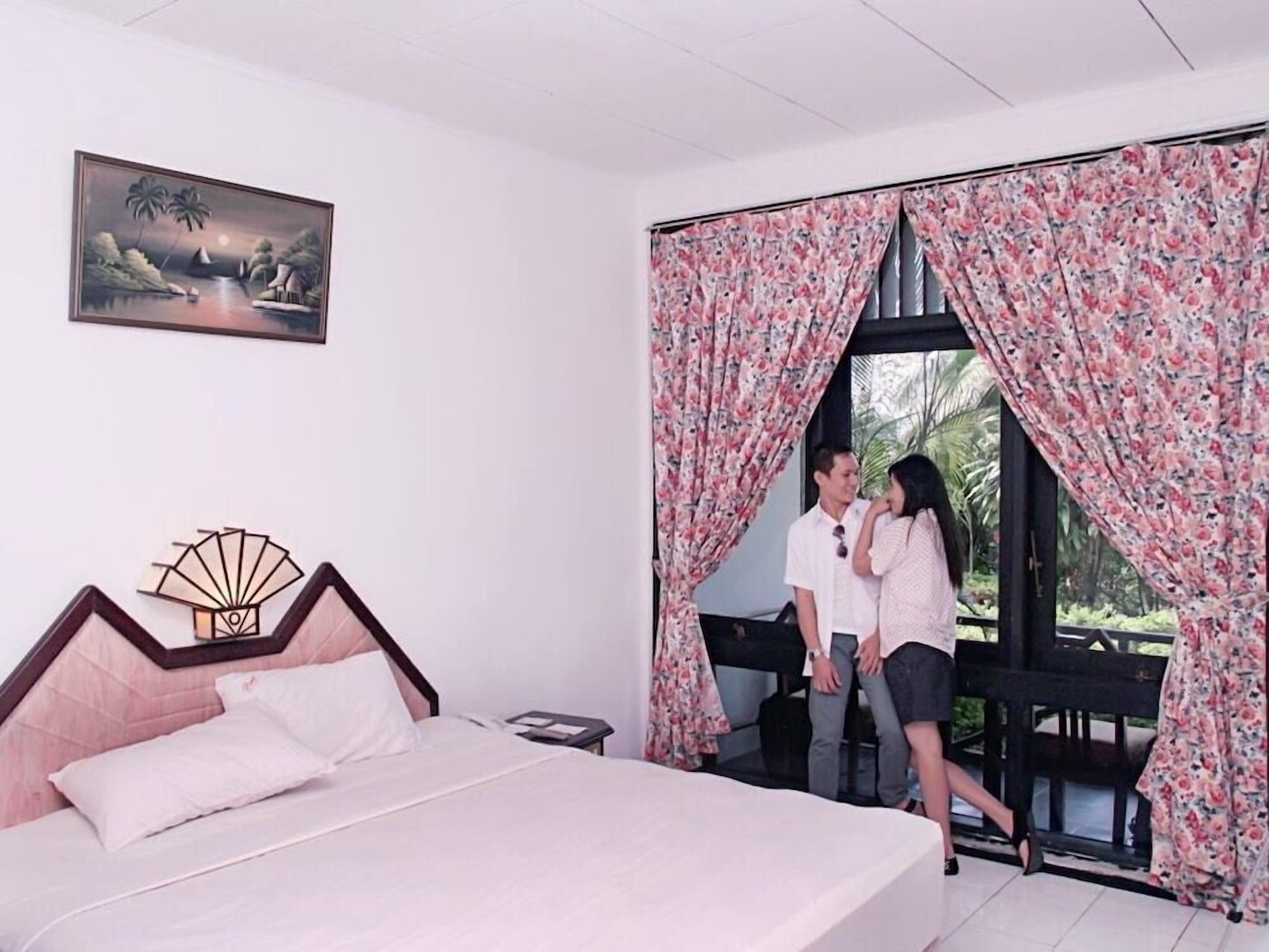 Une chambre à coucher avec un lit, une table de chevet, un tableau et une fenêtre ornée de rideaux à motifs floraux.