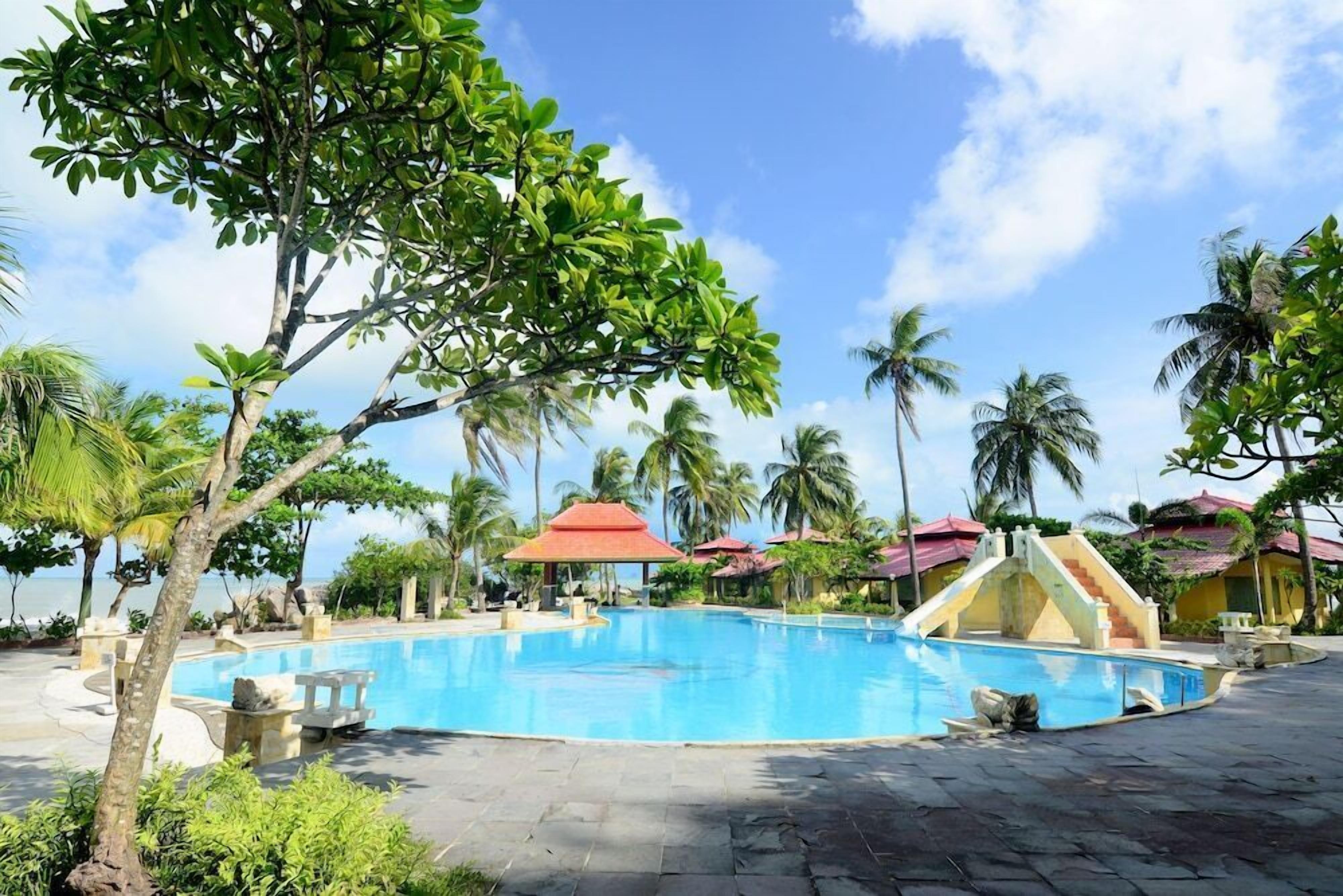 A swimming pool with a red-roofed structure, surrounded by palm trees and a clear sky.