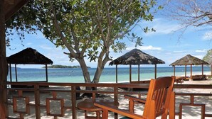 Una terraza de madera con una silla y una mesa, con vista a una playa que tiene sombrillas de paja y una vista despejada del océano.