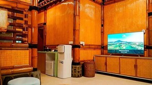 Una habitación con paredes de madera, un televisor mostrando un paisaje, un refrigerador pequeño y un cesto de basura.