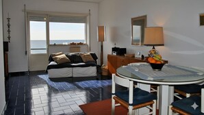 Una stanza con un letto, un tavolo da pranzo con sedie, una ventola a soffitto e vista sul mare attraverso una finestra.
