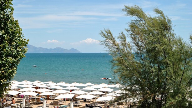 Una spiaggia con file di ombrelloni e lettini, vista libera sul mare e montagne sullo sfondo.