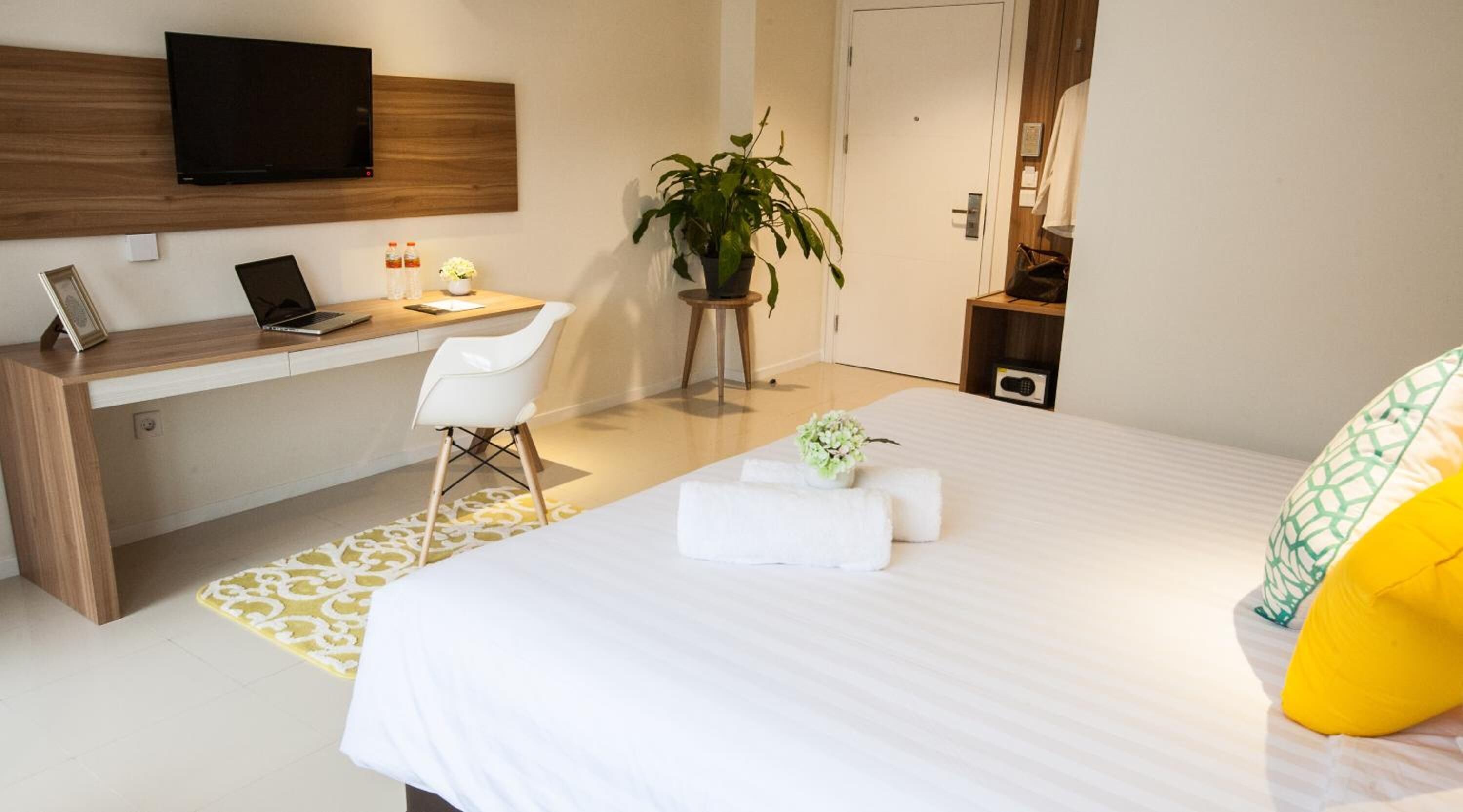A hotel room with a bed, a desk with a laptop, a chair, a potted plant, and a television on the wall.