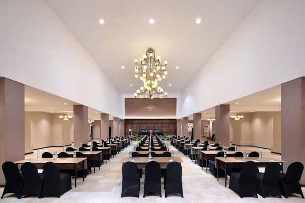 Een congreszaal met lange tafels en stoelen, een kroonluchter en een podium aan de voorzijde.