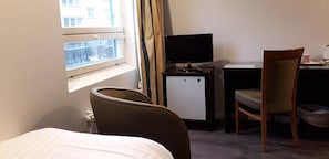 Hotellihuone, jossa on sänky, työpöytä tietokoneella, tuoli ja ikkuna, jossa on verhot.