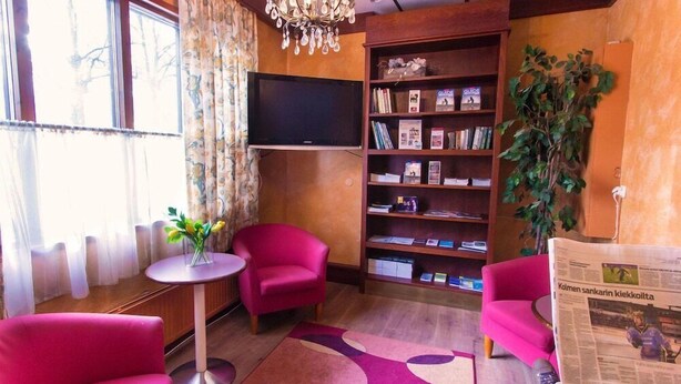 A cozy room with a flat-screen TV, a bookshelf, two pink armchairs, a small table with a vase, and a large plant. - Kauppahotelli Lahti (Lahti)