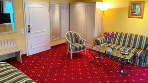 A hotel room with a red carpet, striped sofa, armchair, and a glass coffee table. There are wardrobes and a television on a cabinet. - Woertz Schlosschen Zur Weinsteige (Stuttgart)