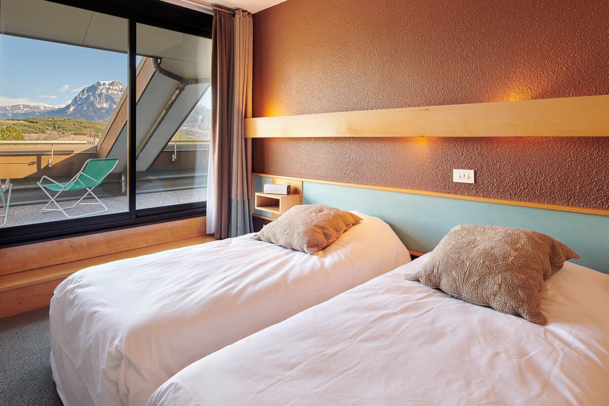 Habitación de hotel con dos camas, una ventana que ofrece vistas a las montañas, una silla verde y un estante de madera.