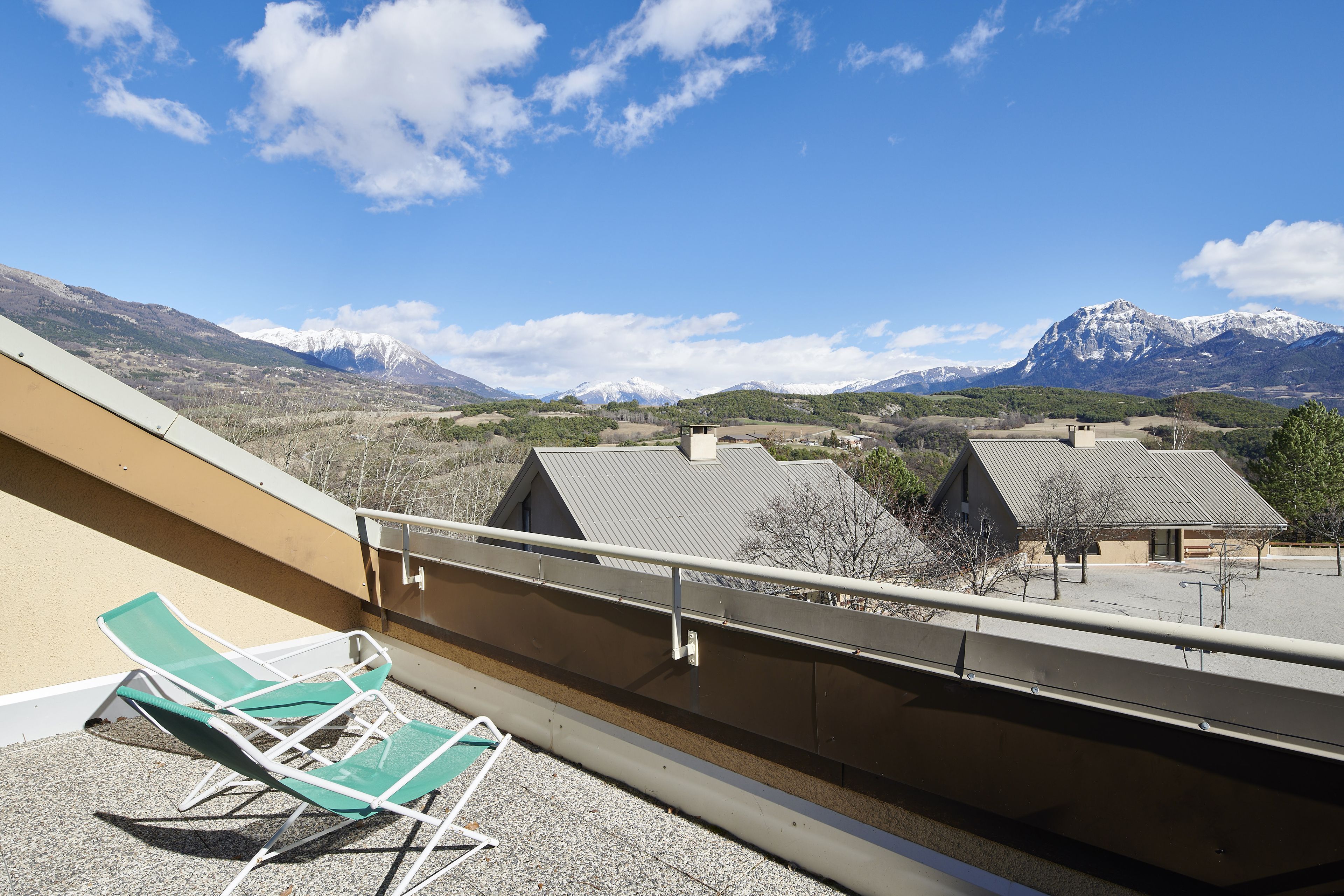 A balcony with two green lounge chairs, a view of snow-capped mountains, and a clear blue sky.