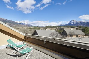 Un balcón con dos sillones verdes, vistas a montañas nevadas y un cielo azul despejado.
