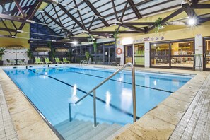 Ein Hallenbad mit Glasdecke, Liegestühlen und im Hintergrund ein Fitnessbereich, der durch Glastüren sichtbar ist.